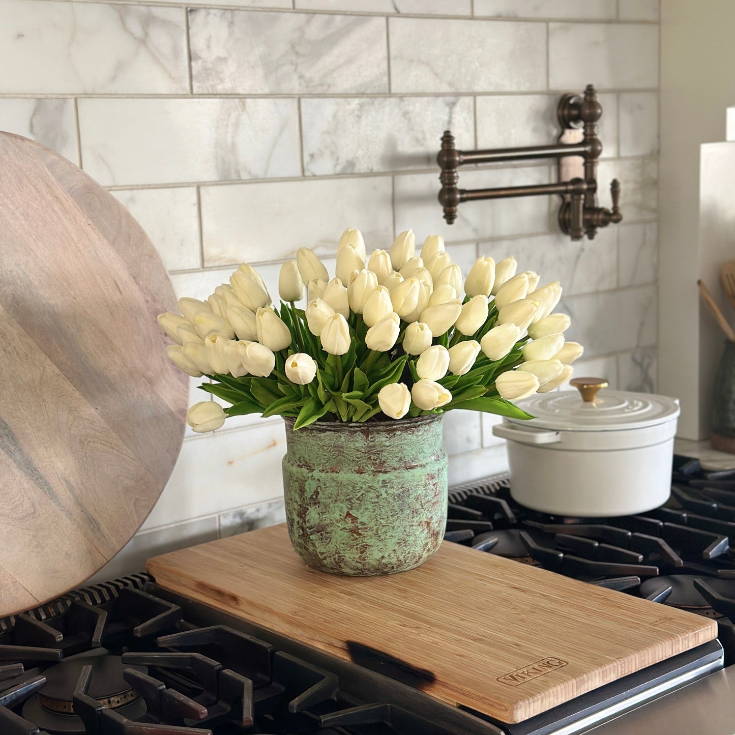 White tulips in a vase on a wooden cutting board over a stove with a tiled wall background