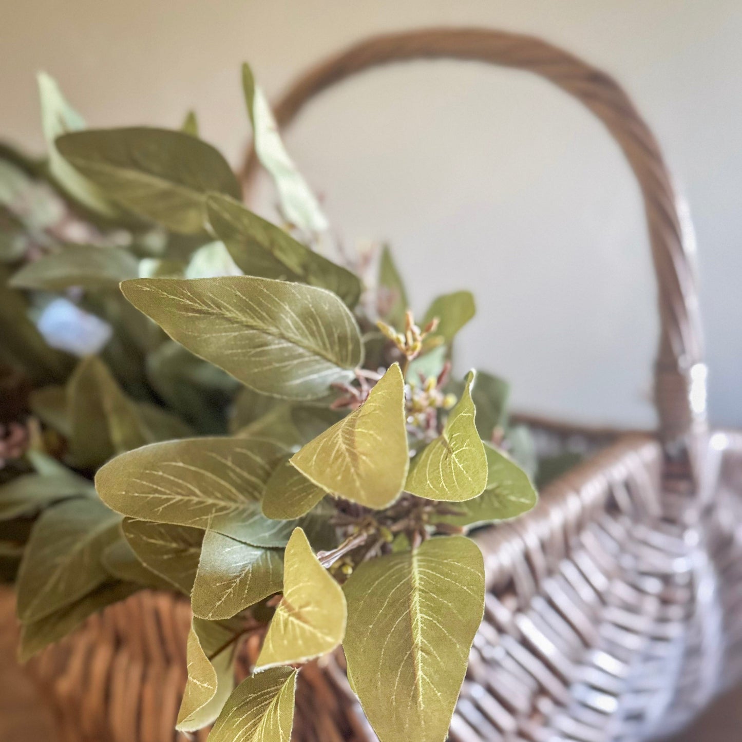 A 23 inch seeded eucalyptus spray with olive green leaves in a wicker basket, depicting artificial greenery for home decor.