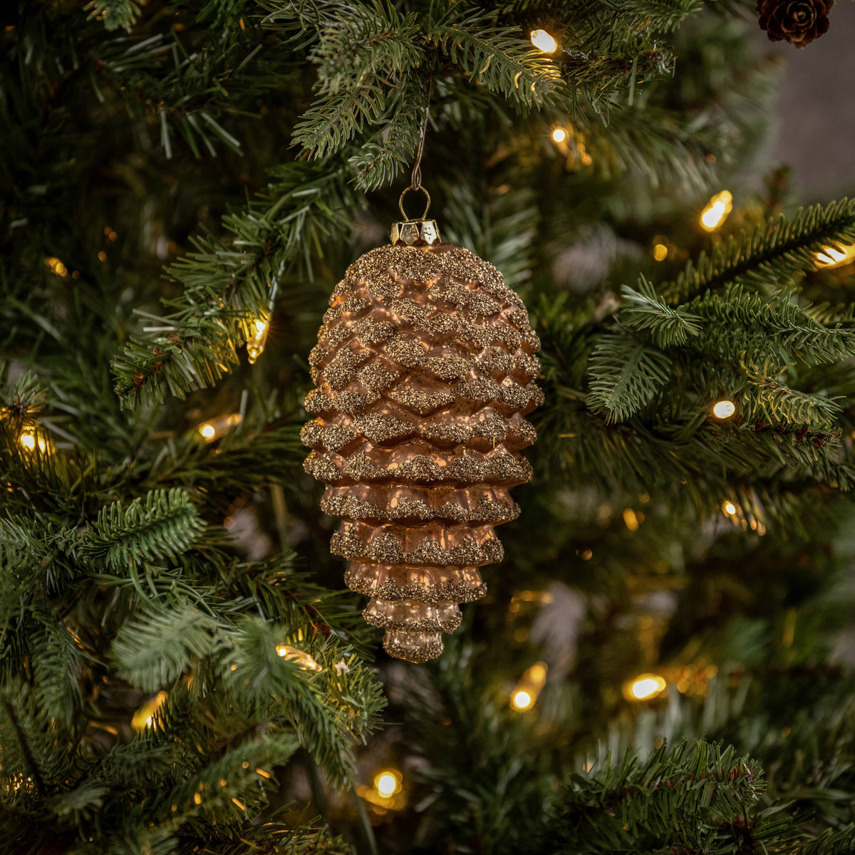 A gold dusted bronze pinecone ornament hanging on a Christmas tree with lights and greenery in the background.