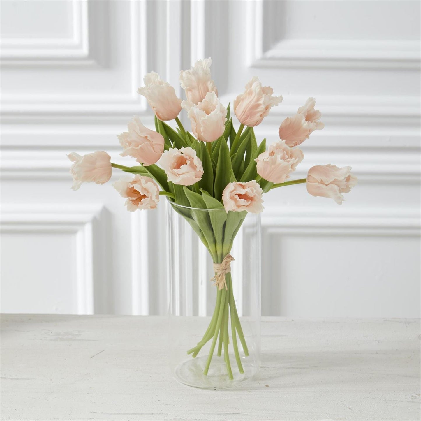 Bouquet of light peach tulips in a clear vase on a white surface with a white paneled wall background.