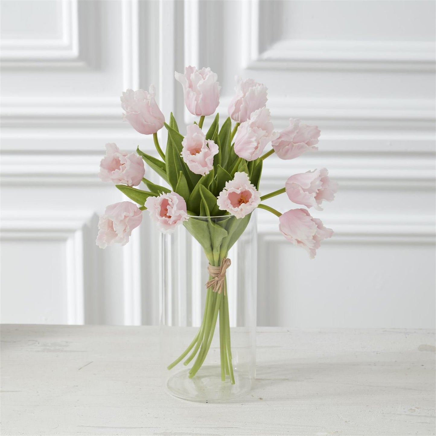 Bouquet of pink tulips in a clear vase on a white surface with a white paneled wall background.