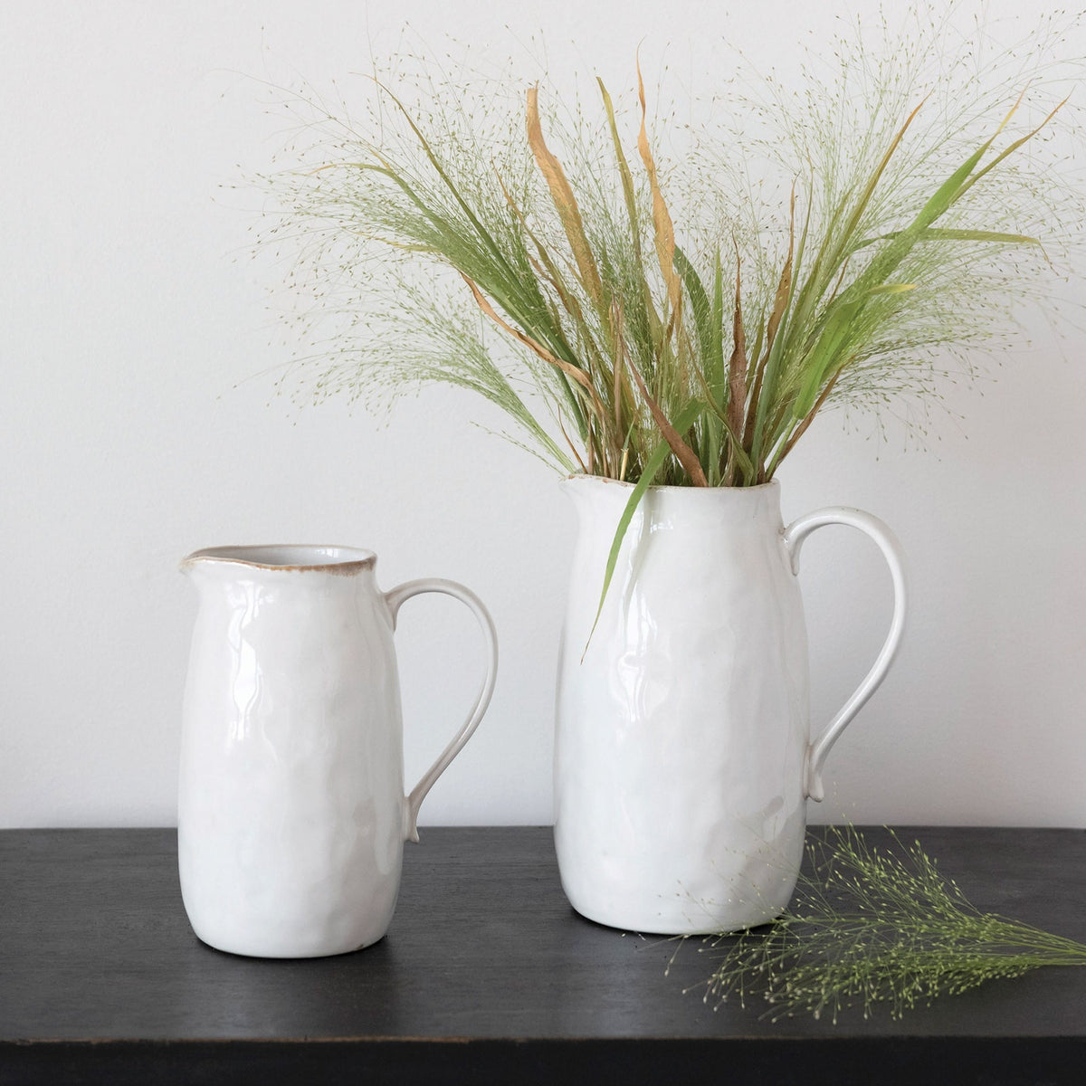Two white ceramic pitchers with plants on a dark surface against a light background