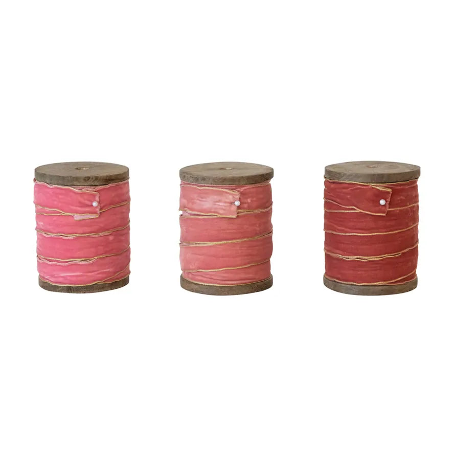 Three wooden spools of velvet ribbon with metallic edges, in shades of pink and red, displayed against a neutral background.