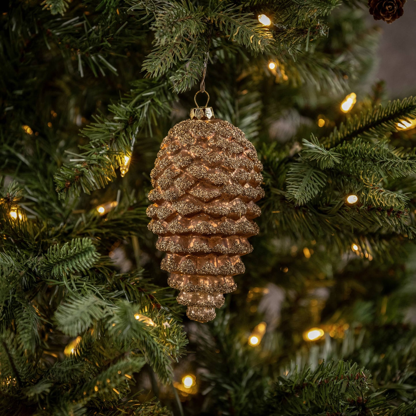 A gold dusted bronze pinecone ornament hanging on a Christmas tree with lights and greenery in the background.