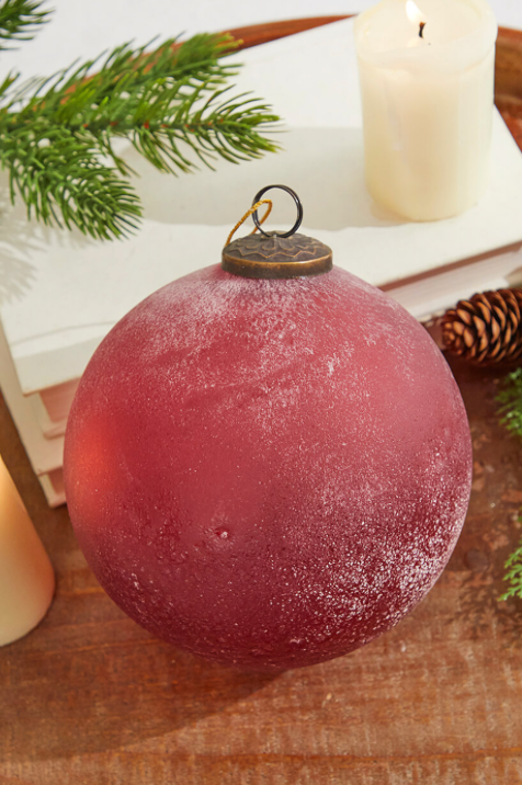A red frosted ruby glass ornament is displayed against a backdrop of Christmas decor, including greenery and a white candle.
