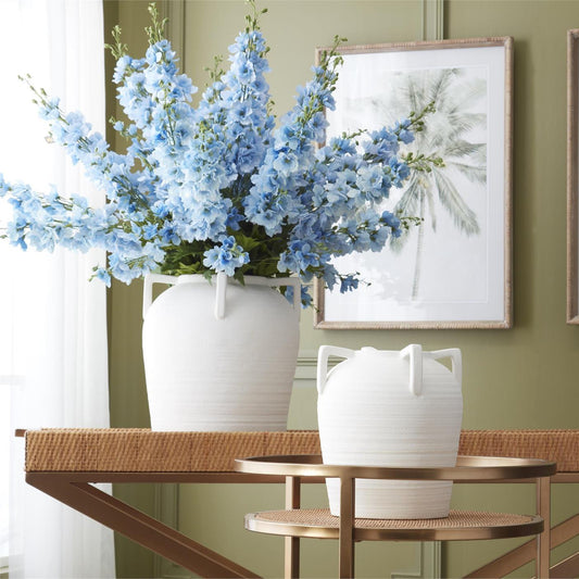 White vases with blue delphiniums on a wooden table against a green wall with framed artwork.