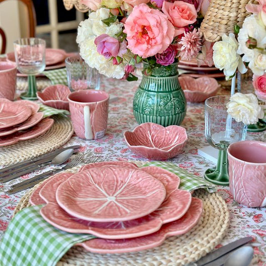 Decorative table setting with pink floral dishes, green vase, and flowers on a patterned tablecloth.