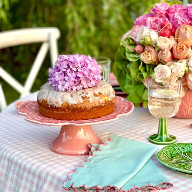 Table setting with a cake, flowers, and glasses on a checkered tablecloth outdoors.