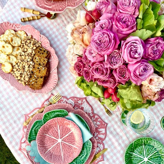 Table setting with pink floral arrangement, plates with food, and decorative items on a checkered tablecloth.