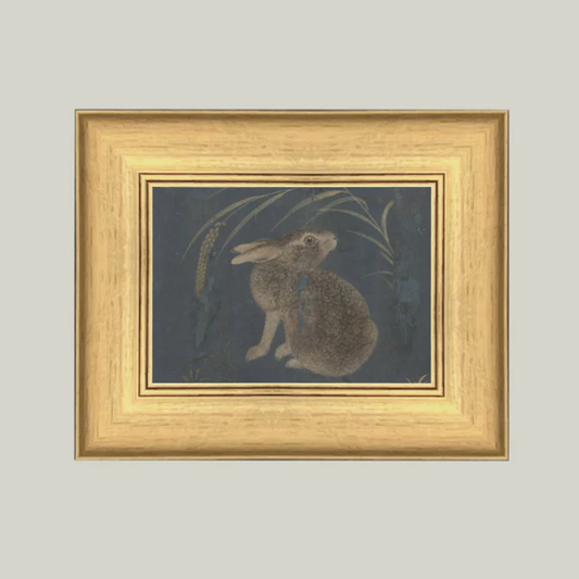 A framed vintage artwork depicting a brown rabbit on a navy background, surrounded by grass-like elements, with a golden frame.