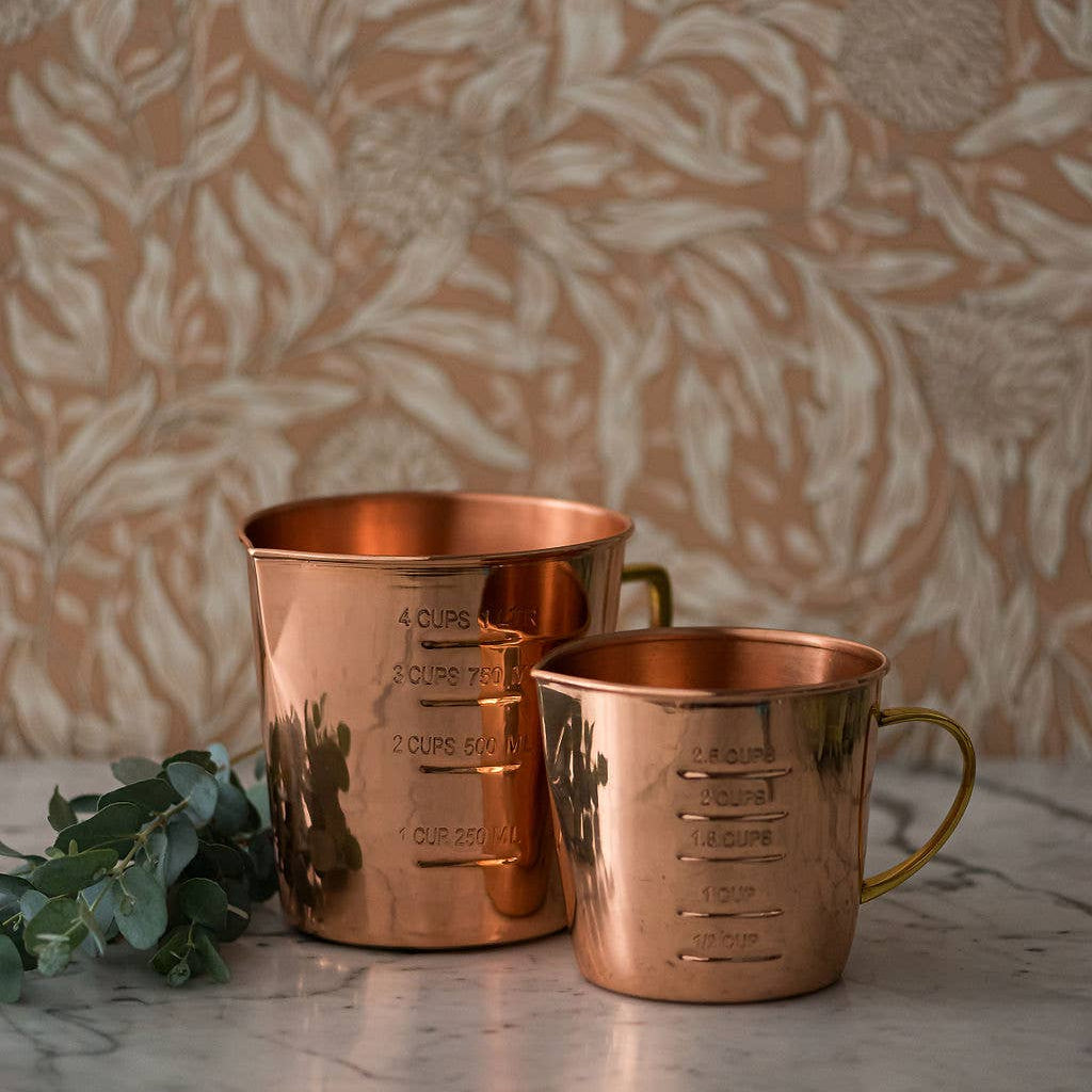 Two copper measuring cups on a marble surface with a floral-patterned wall in the background.
