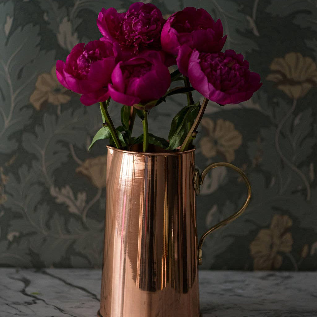 A copper water pitcher with a brass handle, containing pink flowers, placed on a marble surface with a floral pattern in the background.