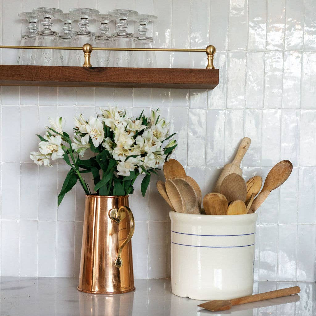 Copper pitcher with white flowers and wooden utensils in a white container on a tiled wall.