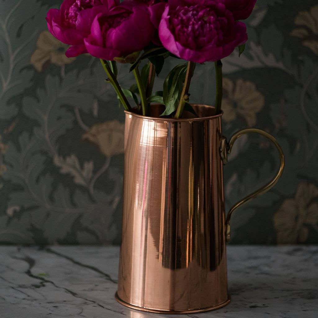 Copper pitcher with purple flowers against a dark floral-patterned wall.