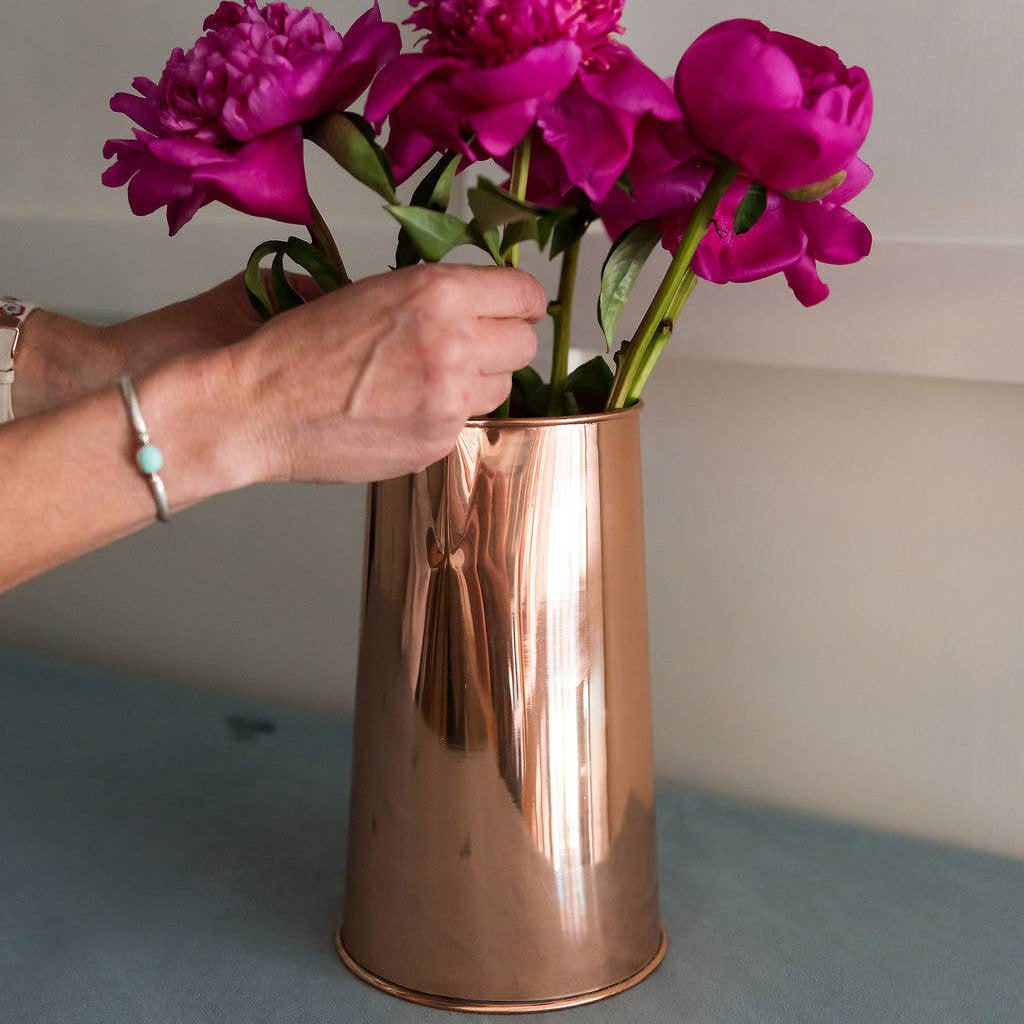 Person arranging purple flowers into a copper vase on a neutral background