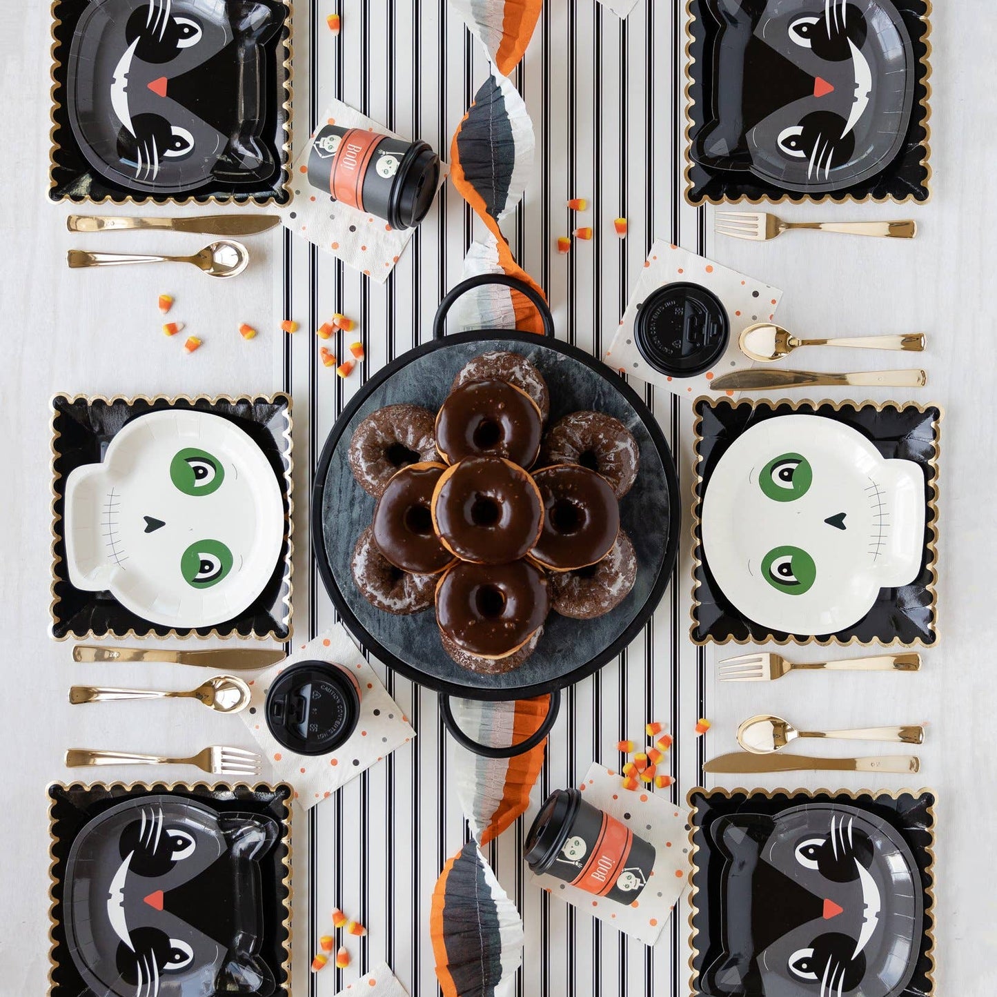 Halloween-themed table setting with black cat plates, donuts, and decorative items on a striped paper table runner.