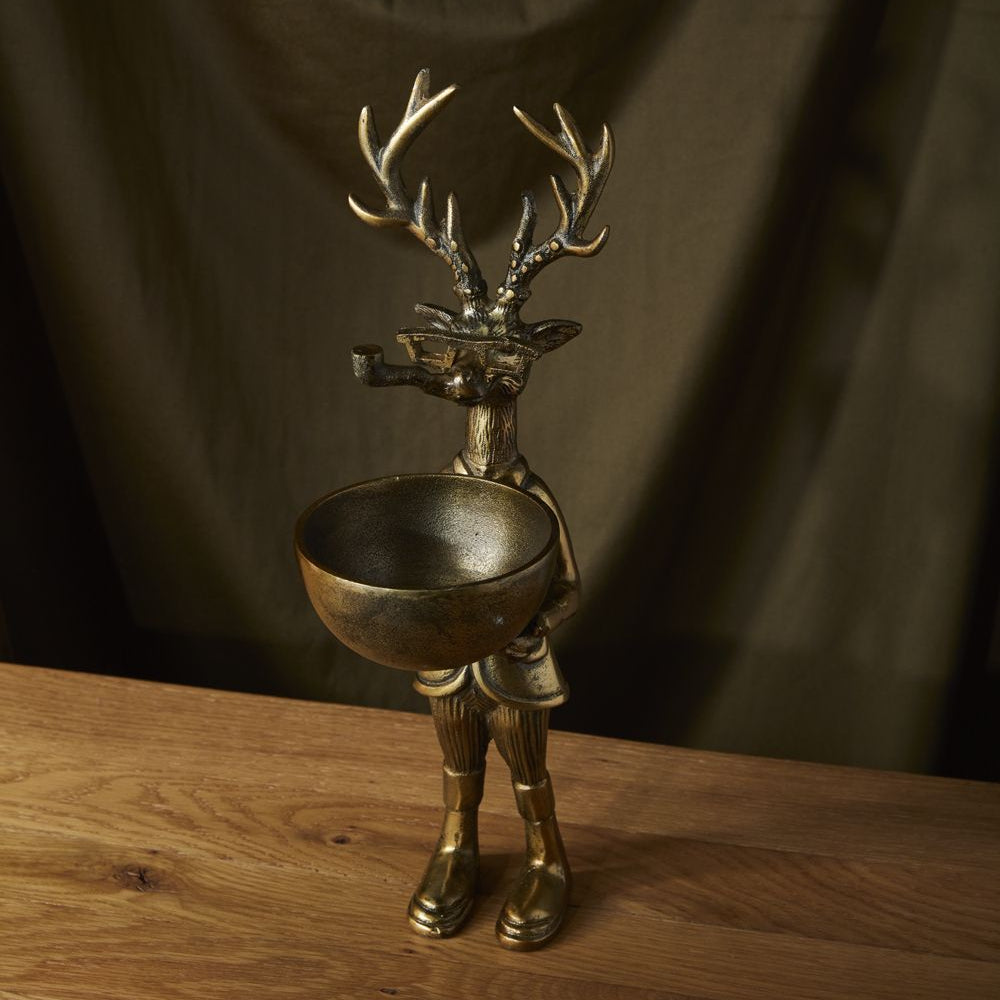A decorative dish stand designed as a figurine of a deer with a dish on its head, set against a dark curtain backdrop and placed on a wooden surface.
