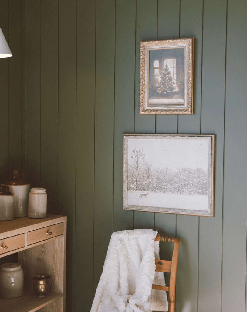 Framed artwork on a green paneled wall with a wooden chair and shelf.