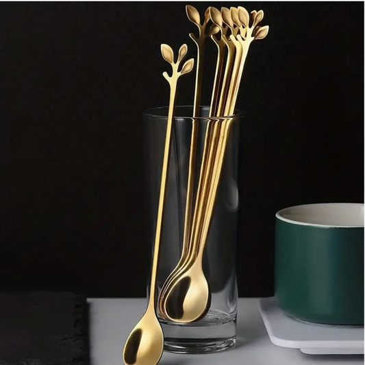 A set of gold-colored, long-handled stainless steel spoons with leaf-shaped handles, displayed inside a clear glass, with a coffee mug and a plate in the background.