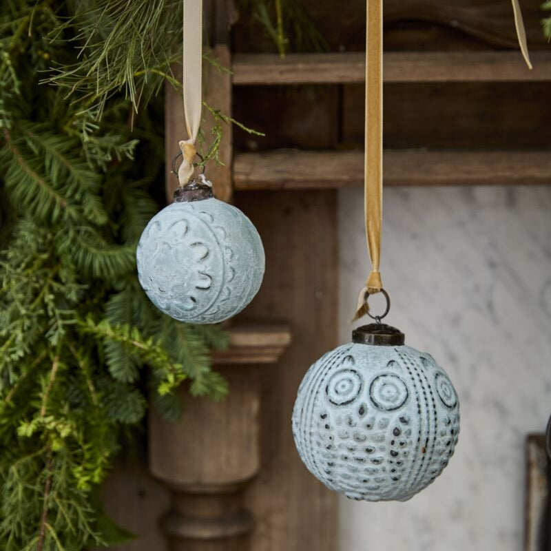 Two light blue glass ornaments with a geometric pattern, hanging from wooden strings, displayed against a backdrop of green foliage and a textured wall.