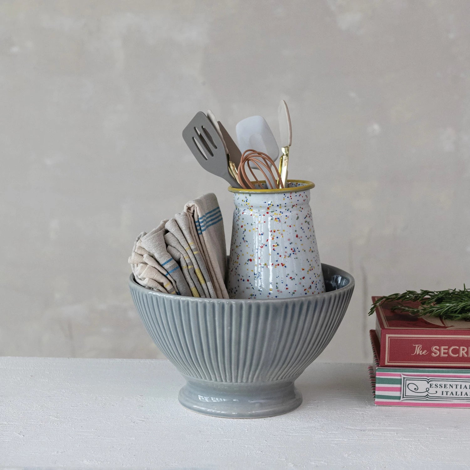 A gray stoneware fluted serving bowl with a reactive glaze, displayed on a table with kitchen utensils inside it, alongside books and a vase.