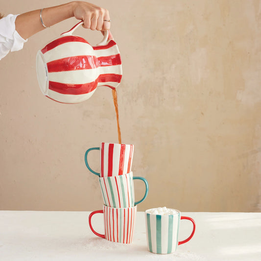Person pouring coffee from a striped red and white pitcher into a stack of colorful striped mugs on a beige background.