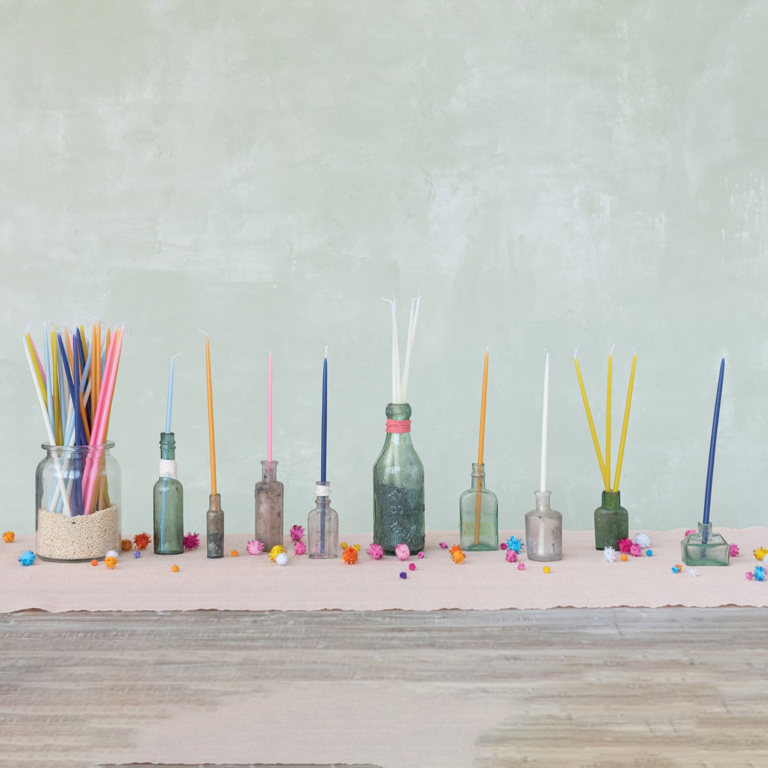 A collection of unscented thin taper candles in various colors including hot pink, displayed in bottles and on a table against a neutral background.