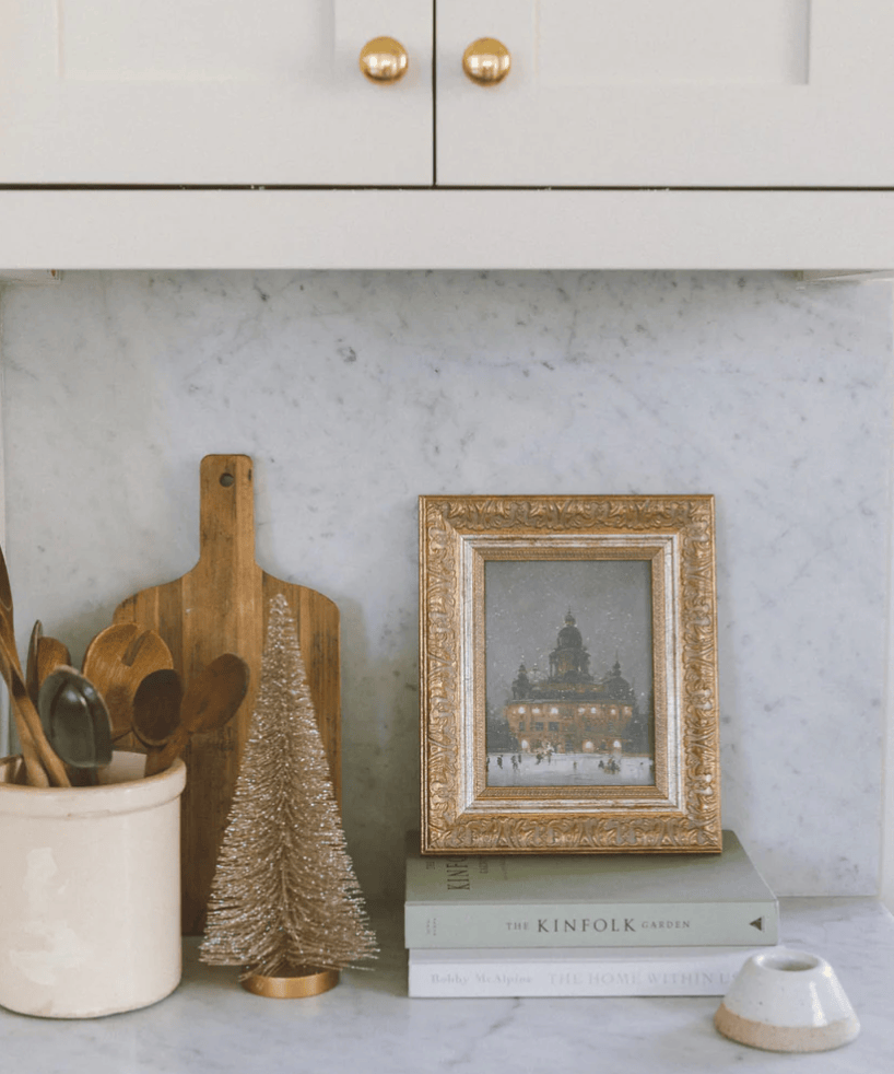 Decorative setup with a framed picture, wooden cutting board, and Christmas tree on kitchen counter.