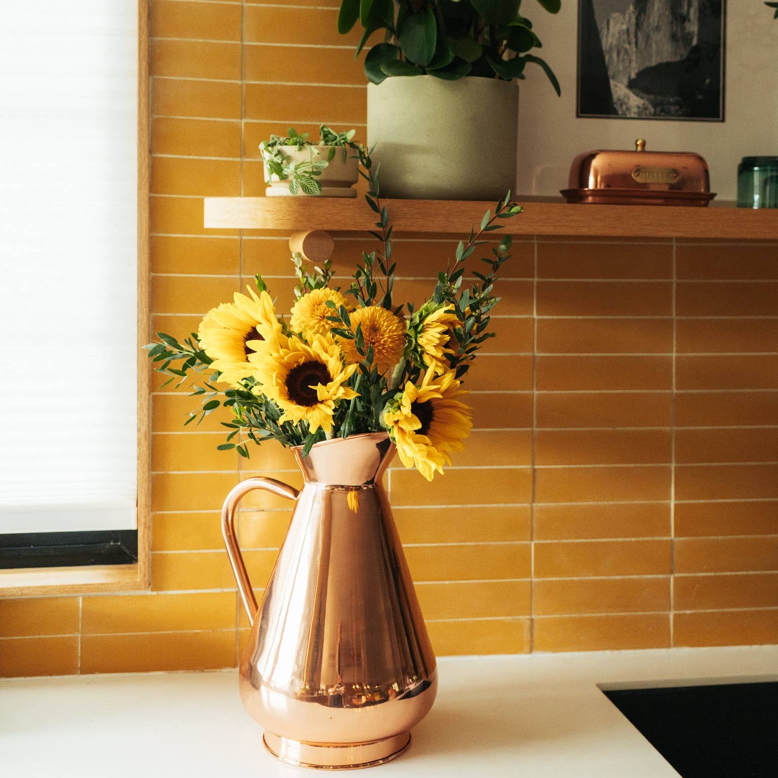 A large copper heirloom vase with a group of sunflowers, placed on a kitchen counter against a yellow tiled wall with decorative plants and a photographic wall shelf in the background.