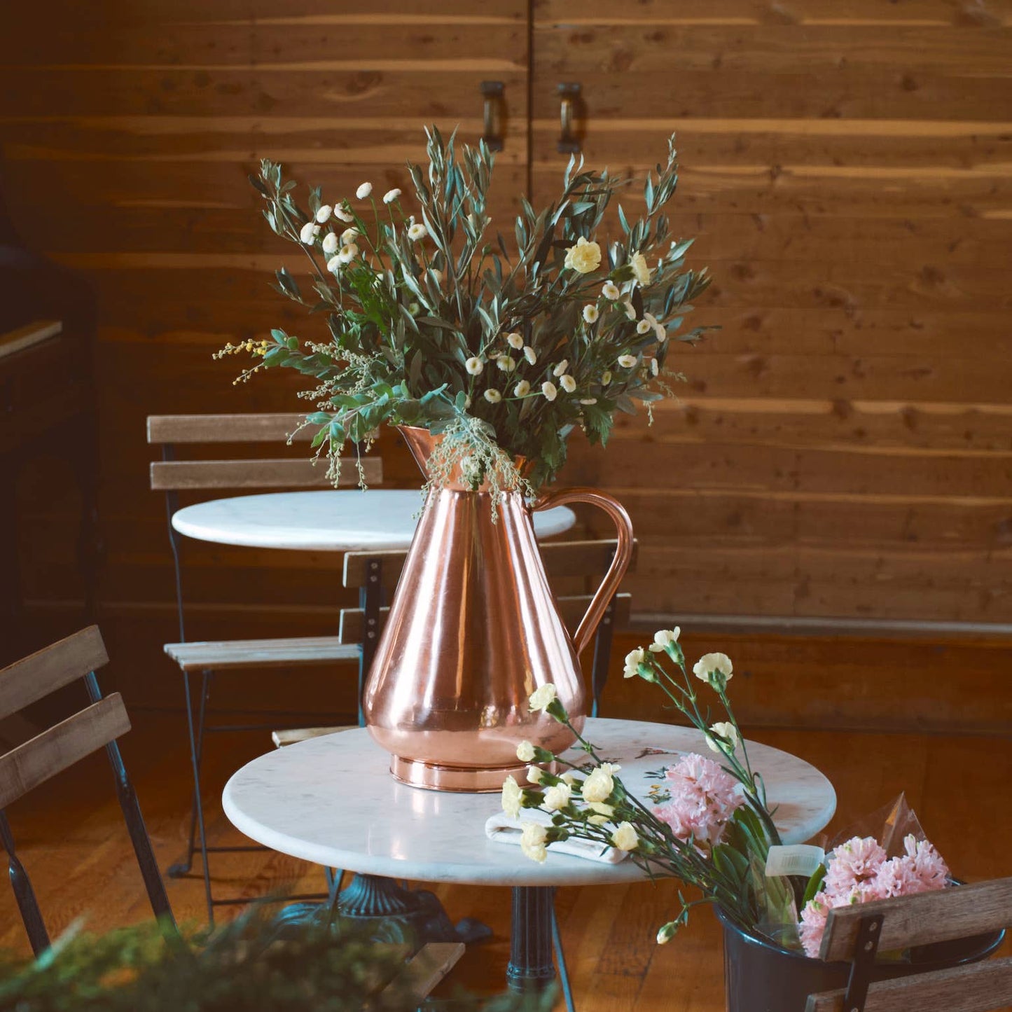 Copper vase with flowers on a table against a wooden wall.