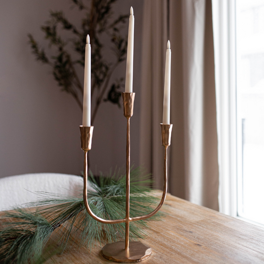 Bronze candelabra with three candles on a wooden table near a window. Luxe B Co. Large 3-Arm Iron Candelabra – Brushed Gold
