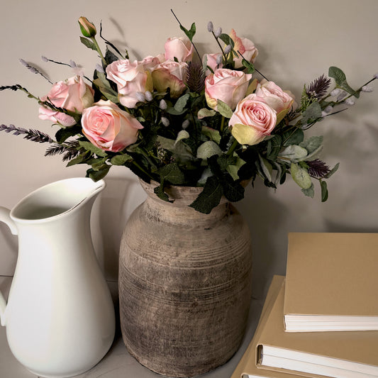 Bouquet of pink roses in a rustic vase on a surface with a white pitcher and beige books.