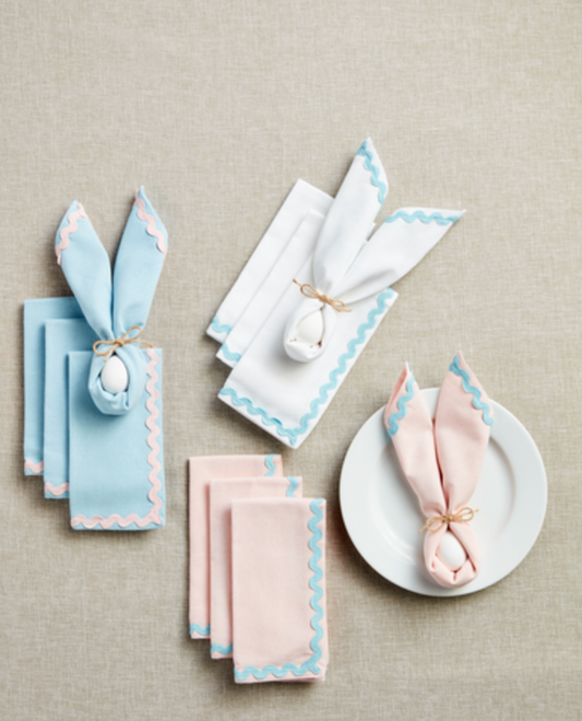 A set of decorative napkins in pink, blue, and white, featuring an ricrac trim, displayed on a table with plates and tied with bows.