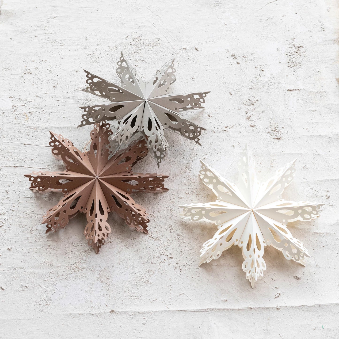 Three paper folding snowflake ornaments in white, silver, and brown colors, displayed on a white surface.