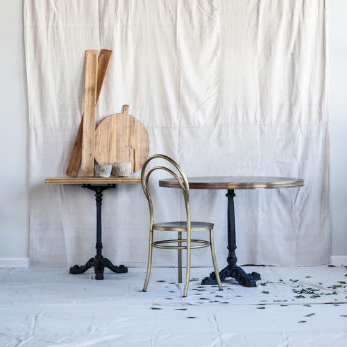 A wooden baguette board leaning against a wall with a round cutting board and a metal chair in the foreground on a white background.