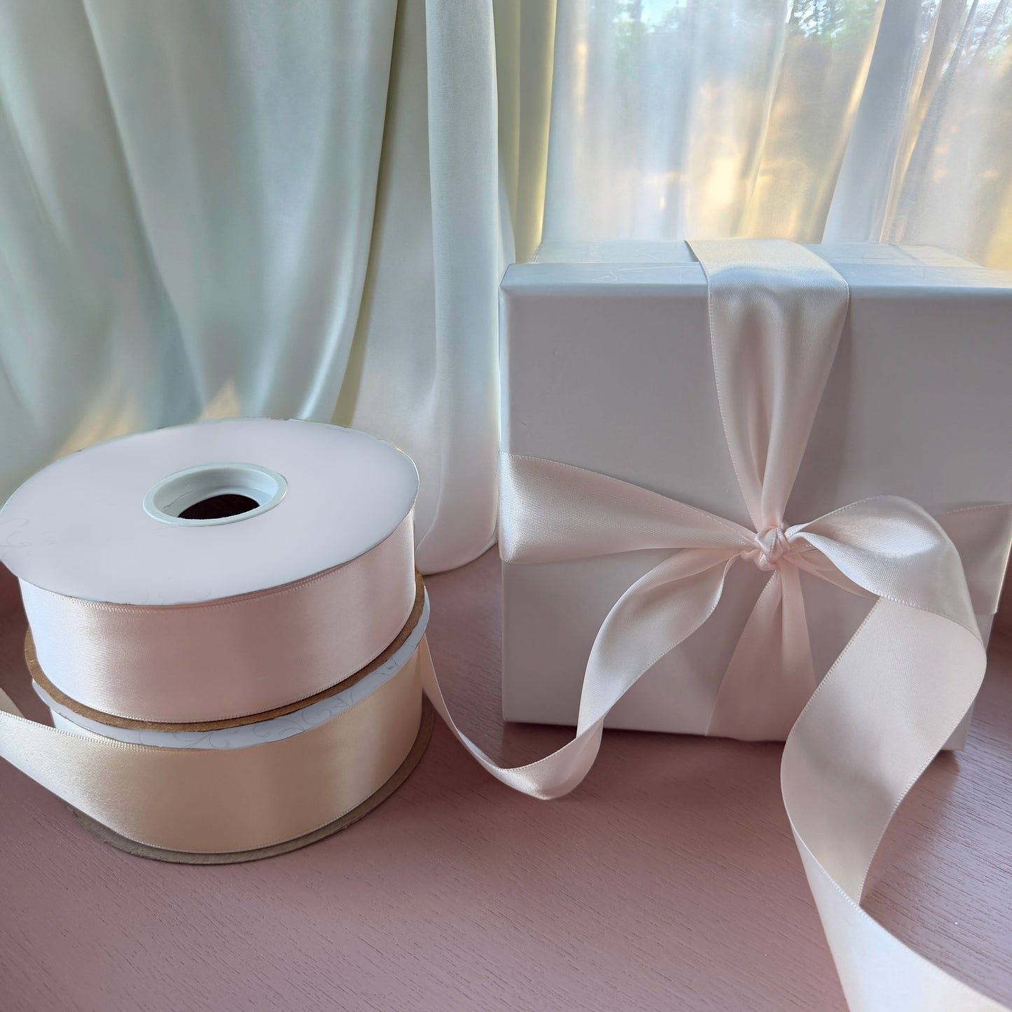 Roll of pink satin ribbon with a white gift box tied with a bow against a light curtain background.