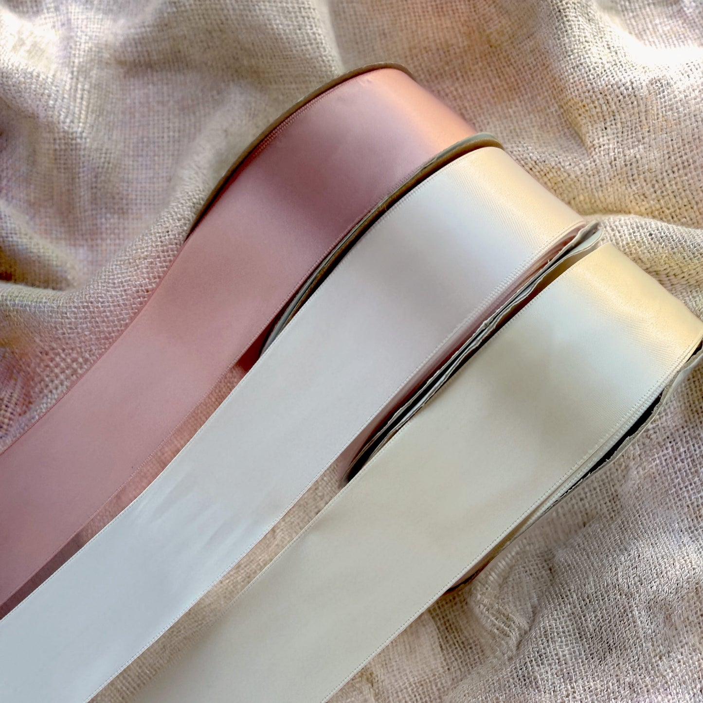 Three rolls of ribbon, two pink and one ivory, on a textured fabric background.
