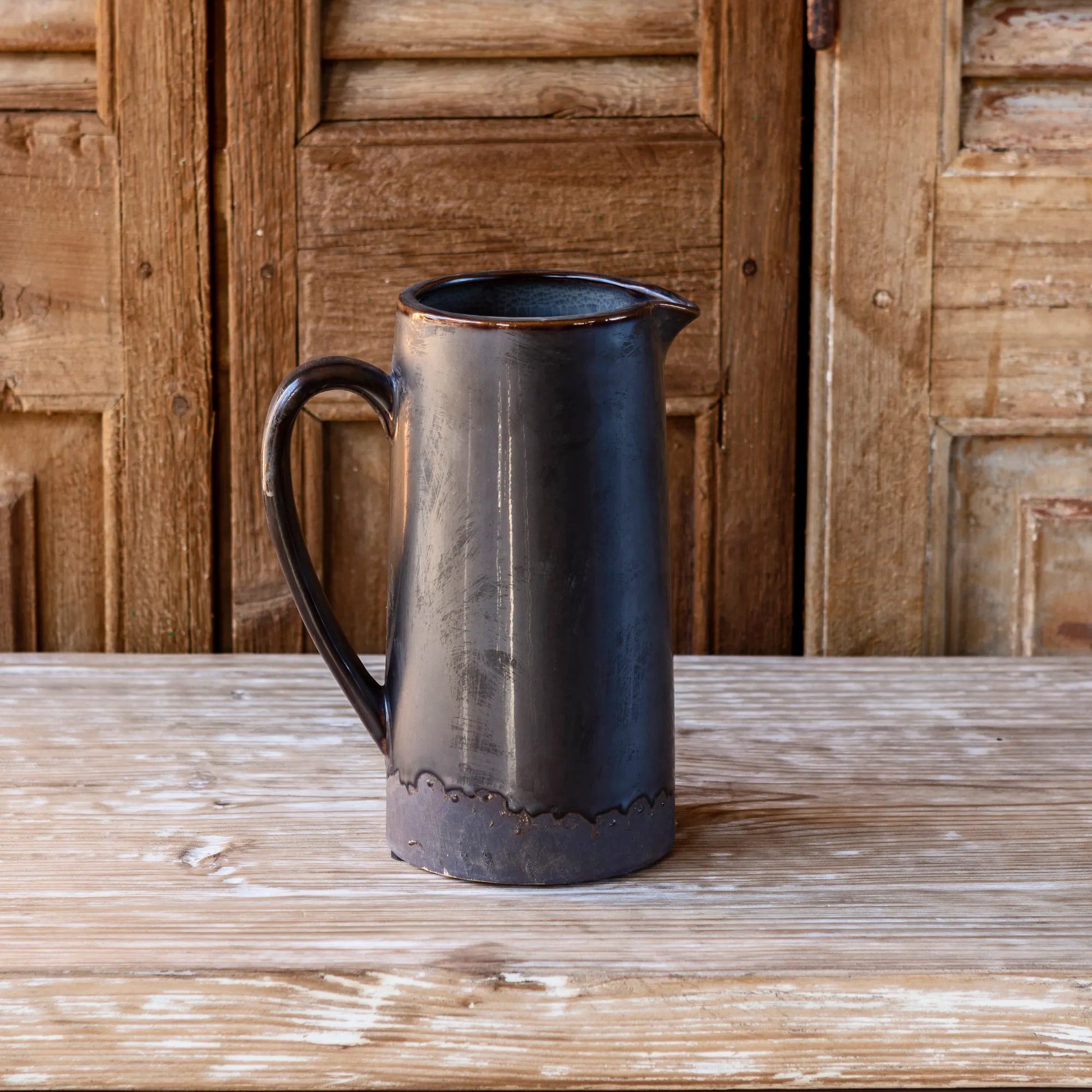 A tall pitcher vase made of glazed pottery with a dark color and a handle, displayed on a wooden surface.