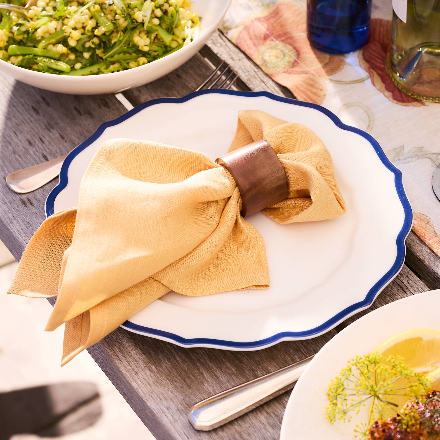 Folded yellow napkin with a wooden napkin ring on a white plate with blue rim, set on a wooden table with a bowl of salad in the background.