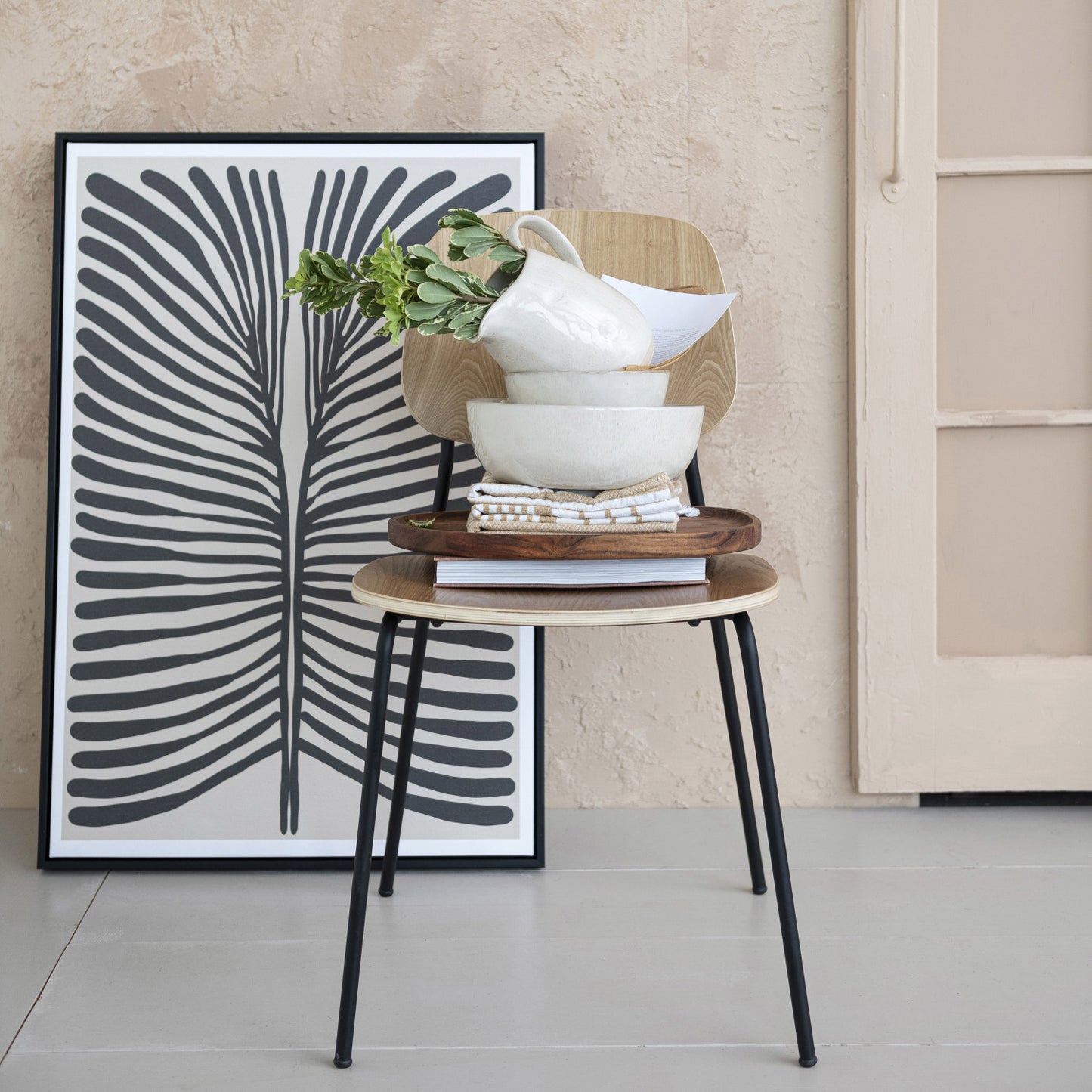 Two wooden stools with a stack of bowls and a plant, against a wall with a black and white abstract art print.