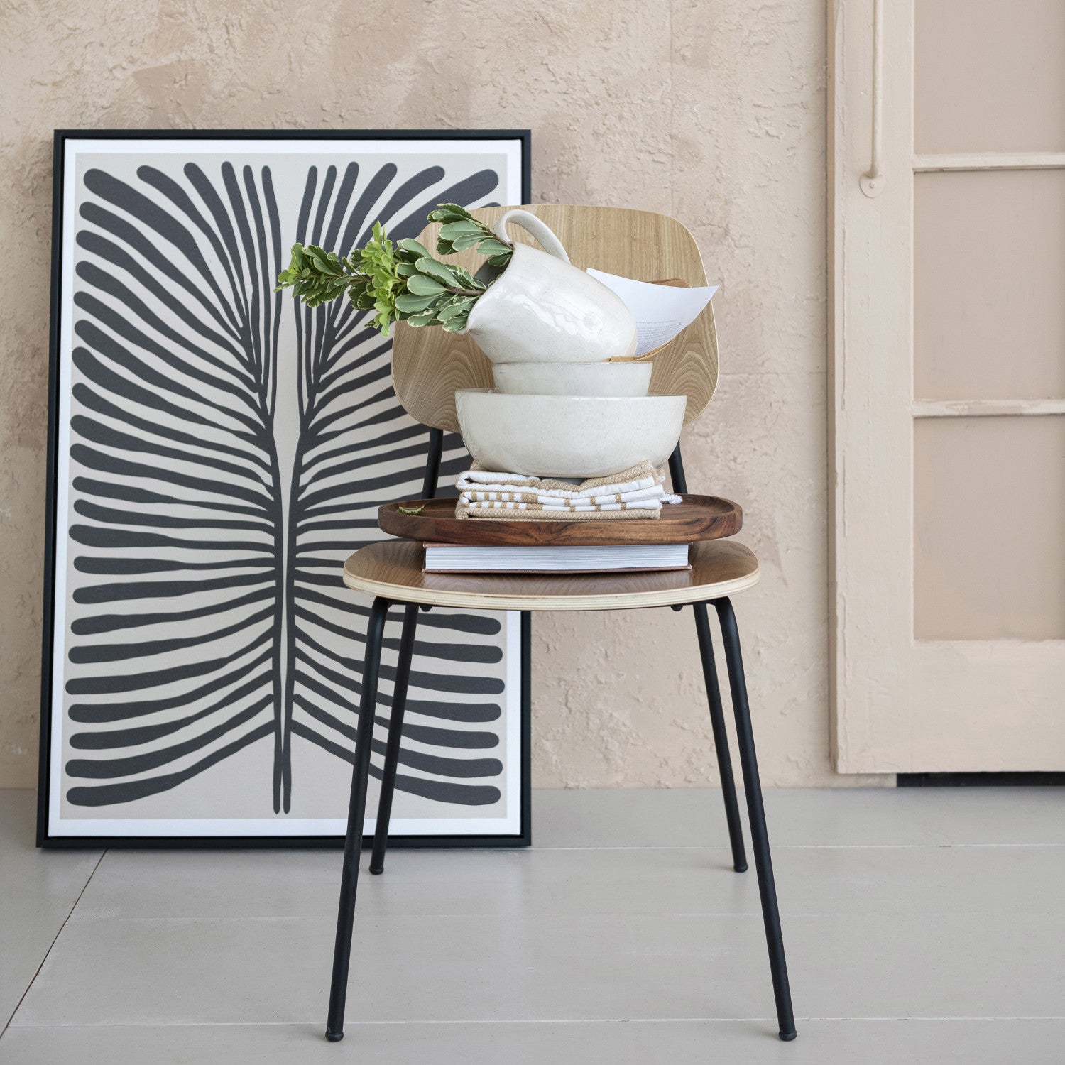 Two wooden stools with a stack of bowls and a plant, against a wall with a black and white abstract art print.