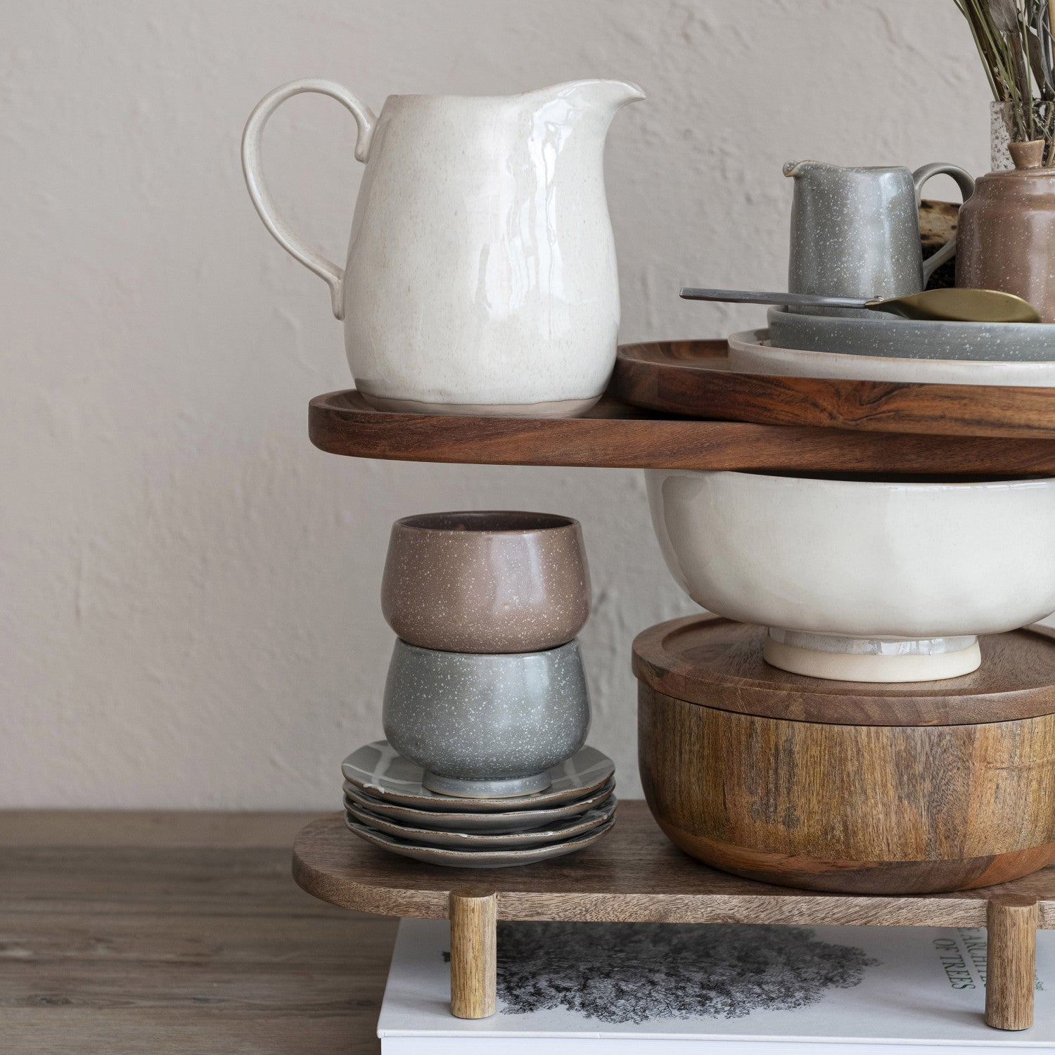 Wooden tiered stand with ceramic items including a pitcher, bowls, and cups on a neutral background.