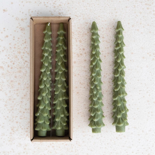 Two green tree-shaped candles next to a box containing more candles on a textured white surface.