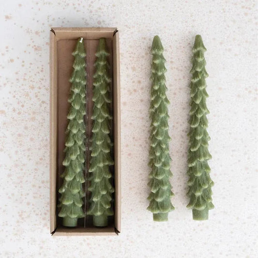 Two green tree-shaped candles next to a box containing more candles on a textured white surface.