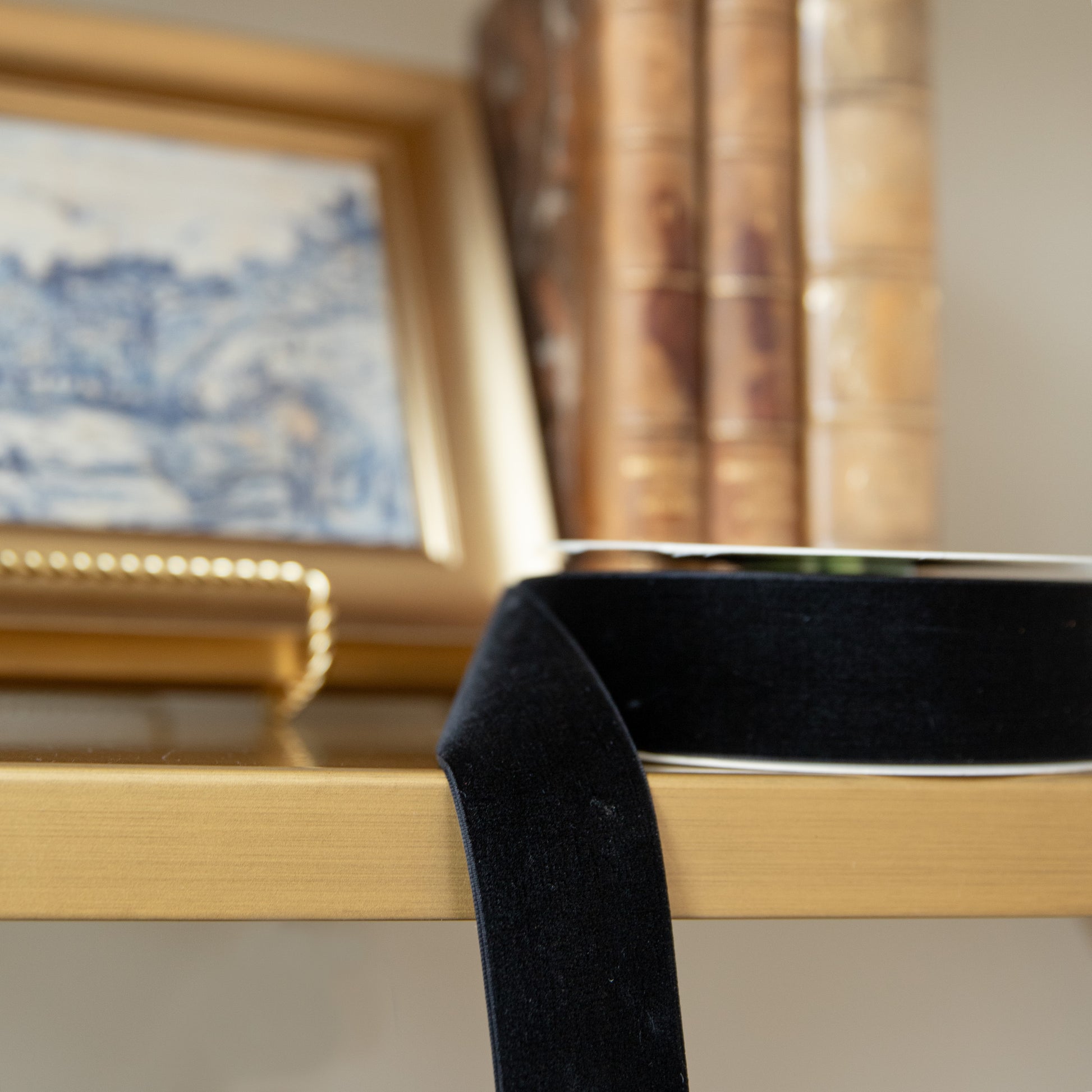 Gold frame with a black ribbon on a surface, with a blurred painting and curtain in the background.