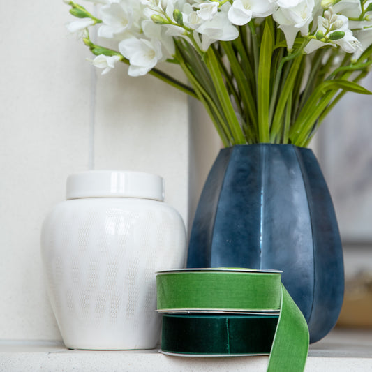 White vase, blue vase with flowers, and green ribbon on a white surface.