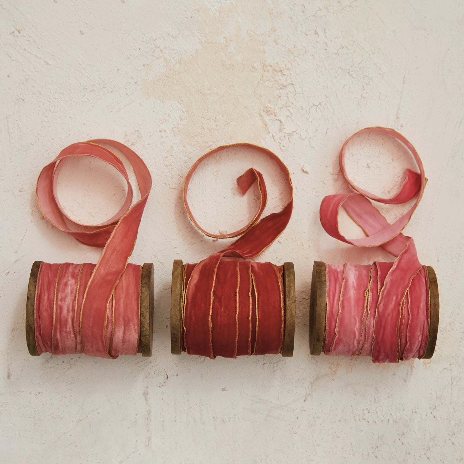 Three wooden spools of velvet ribbon with metallic edges, in shades of pink and red, displayed against a neutral background.