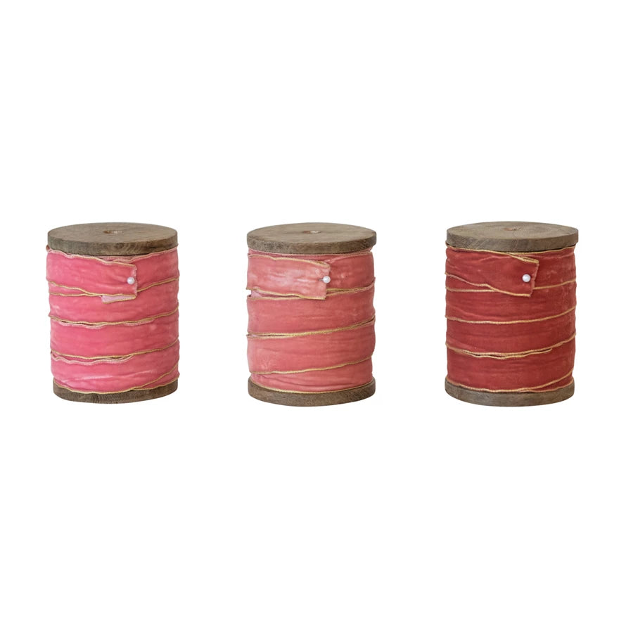 Three wooden spools of velvet ribbon with metallic edges, in shades of pink and red, displayed against a neutral background.