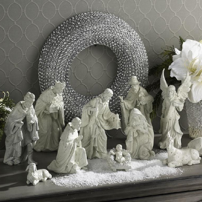 Nativity scene with white figurines on a decorative shelf with a wreath and flowers in the background.