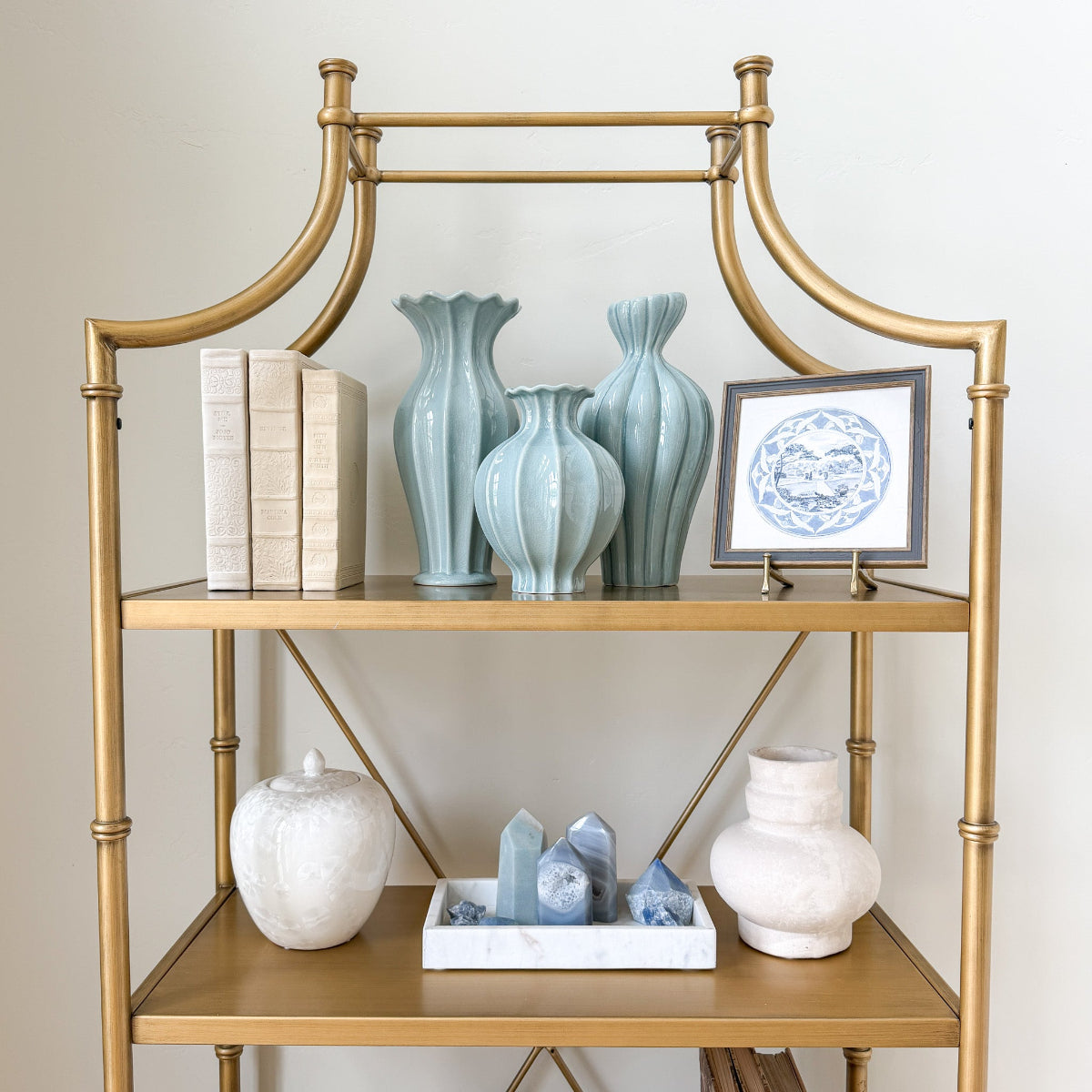 Gold shelf with decorative items including three sizes and styles of light blue vases and a framed picture against a neutral wall.